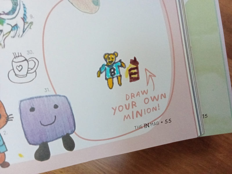A child's hand-drawn and coloured illustration of a golden teddy bear wearing a teal shirt with the letter B, holding what appears to be a bottle, drawn inside a circle prompt in a magazine that says 'Draw your own minion!'