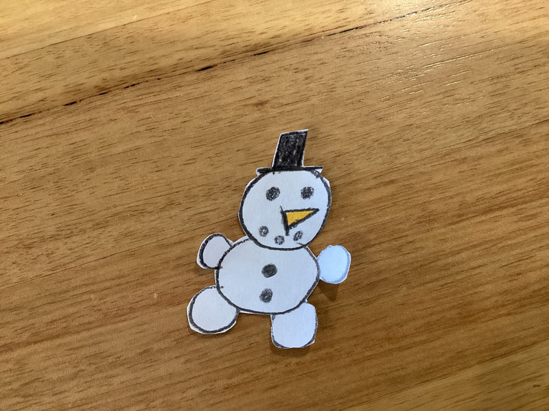 A hand-drawn paper cutout of a cheerful snowman with a black top hat, orange carrot nose, coal eyes, and round arms and legs. The snowman is drawn with pencil and coloured pencil on white paper, and is photographed on a wooden surface.