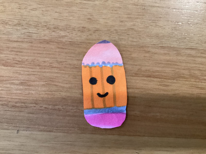 A small oval-shaped paper cutout decorated with orange, pink, and blue stripes, featuring a hand-drawn kawaii-style face with two round black eyes and a small smile. The character is lying on a wooden surface.