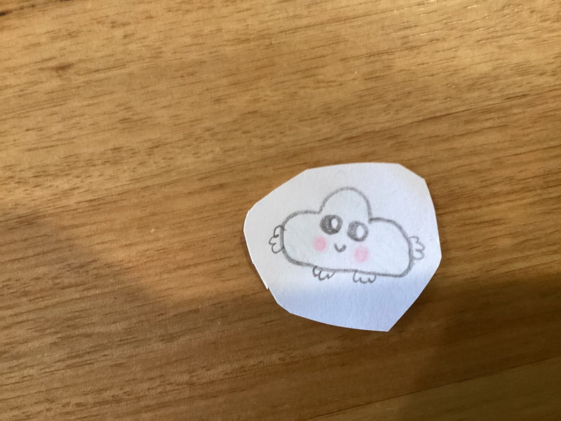 A small hand-drawn kawaii-style cloud character on a cut-out piece of paper, sitting on a wooden surface. The cloud has big round eyes, rosy pink cheeks, a small smile, and tiny wings on its sides.