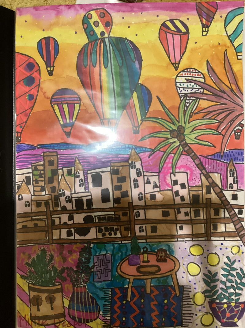 A vibrant, colourful artwork depicting a scene in Cappadocia, Turkey, with many decorated hot air balloons filling a warm orange and pink sunset sky above white buildings and a palm tree. In the foreground, a balcony scene features potted plants, a small table with objects, and patterned textiles.