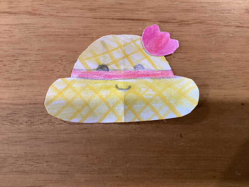 A hand-drawn and cut-out paper craft of a kawaii-style yellow straw hat with a pink band and a pink flower on top. The hat has a cute smiling face drawn on it and is decorated with a yellow grid pattern.