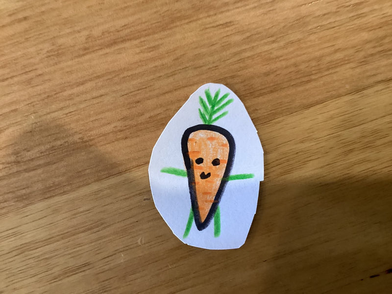 A hand-drawn cartoon carrot character with a smiley face, green leafy top, and small stick arms, drawn in orange and black marker on a cut-out piece of white paper, resting on a wooden surface.