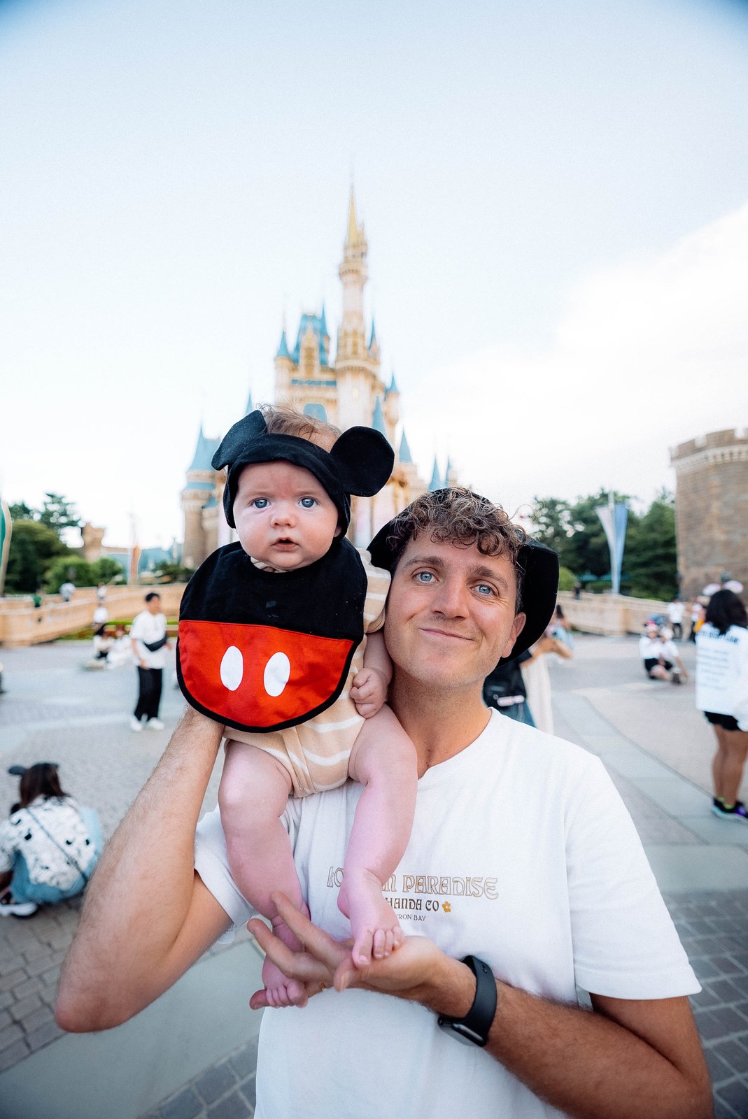 A dad holds a baby dressed as Mickey Mouse in front of the Disney castle.
