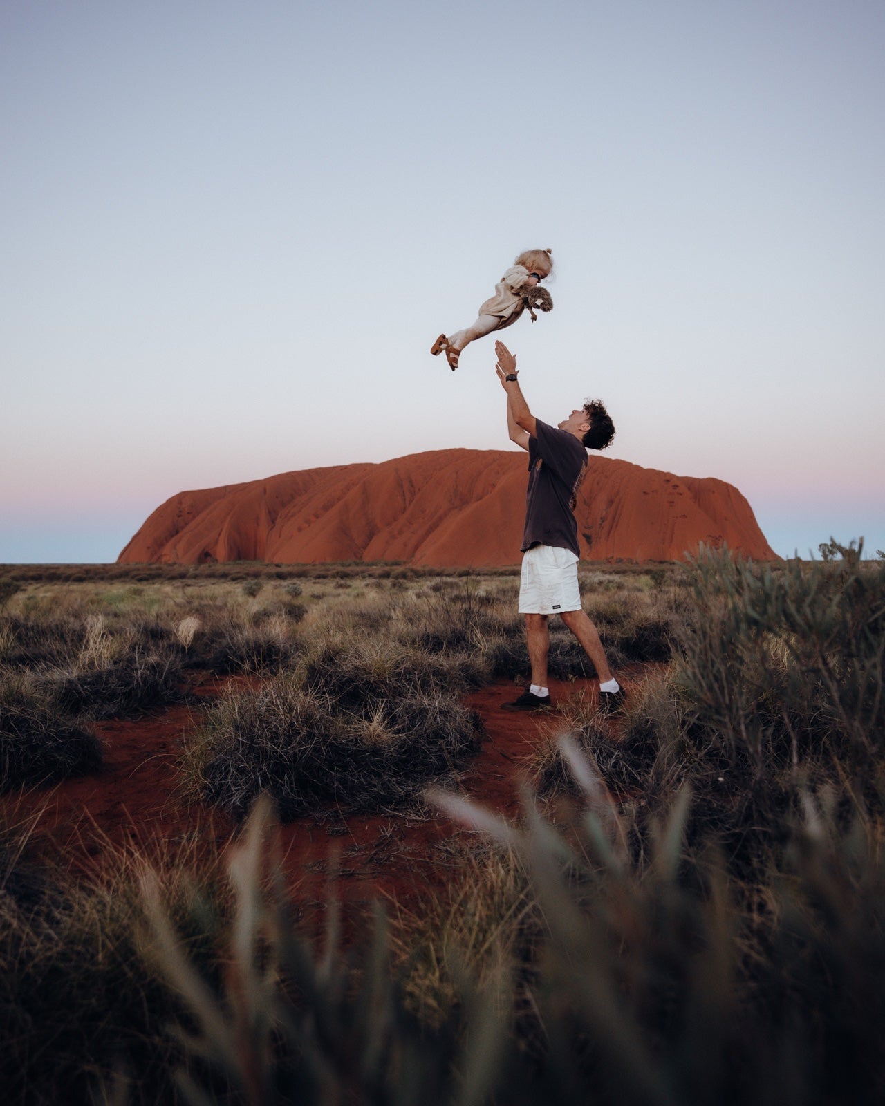 A dad lifts one of his kids into the air at sunset in front of Uluru.