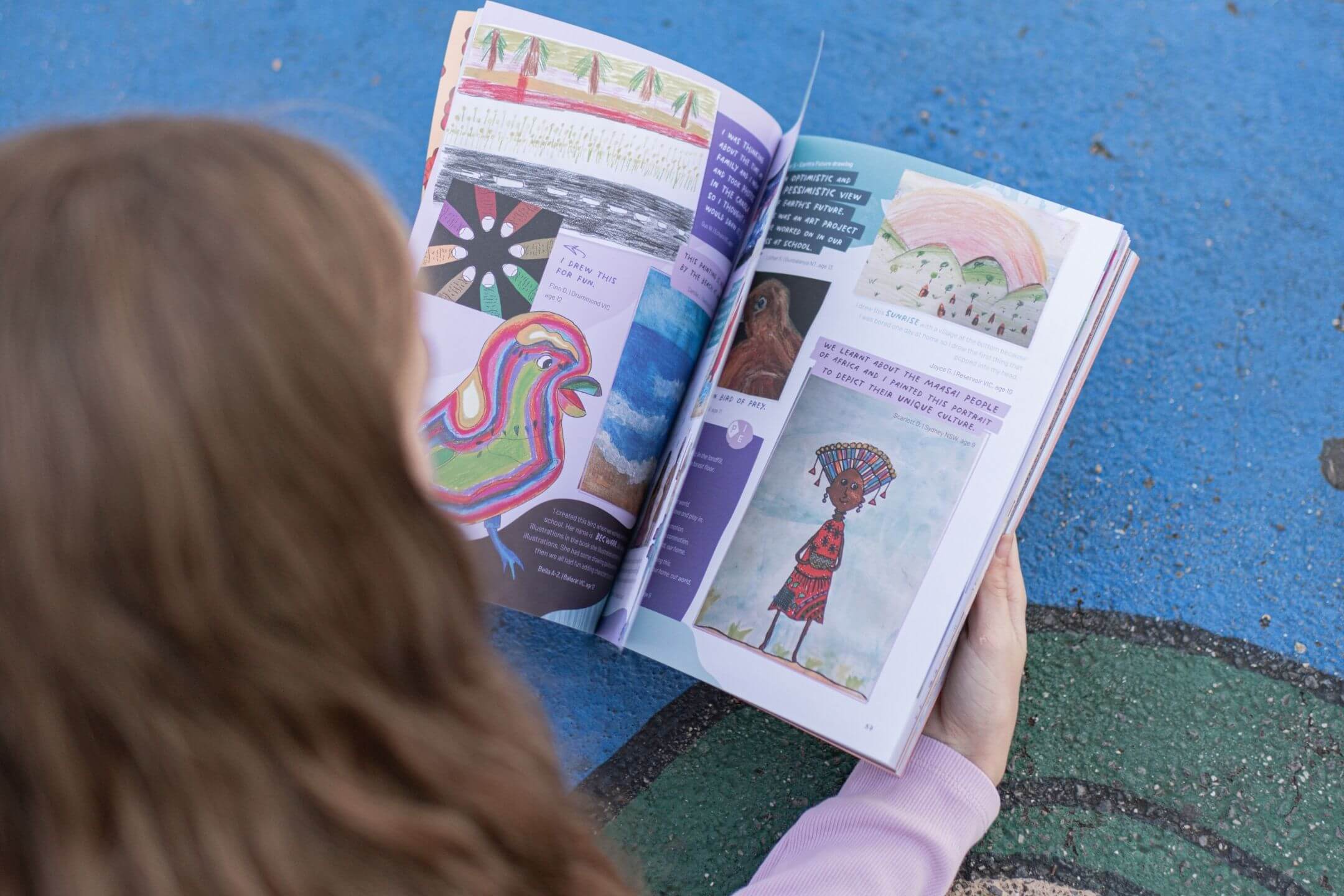 Over-the-shoulder view of a reader looking through an open issue of theINmag, its pages full of colourful kid creations including a bright bird painting and a hand-painted portrait.